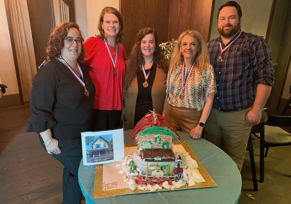 A group of people posing in front of a gingerbread house.
