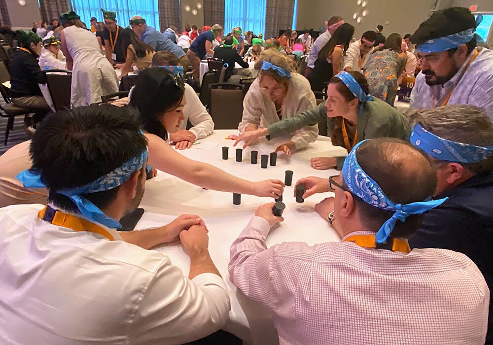 A group of people wearing blue bandanas sit around a table engaged in a team activity involving stacking small cylindrical objects. Other groups can be seen at tables in the background.