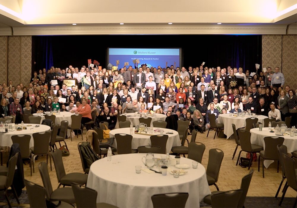 A large group of people pose for a photo in a conference hall with round tables and an overhead screen in the background.