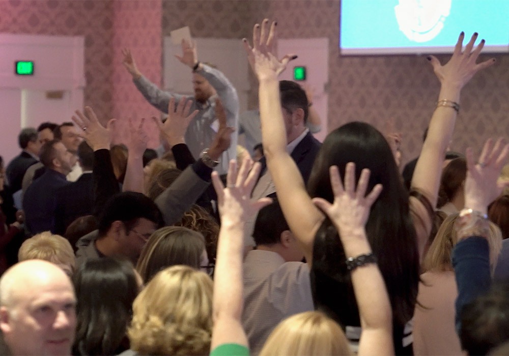 A group of people in a conference room with their hands raised.
