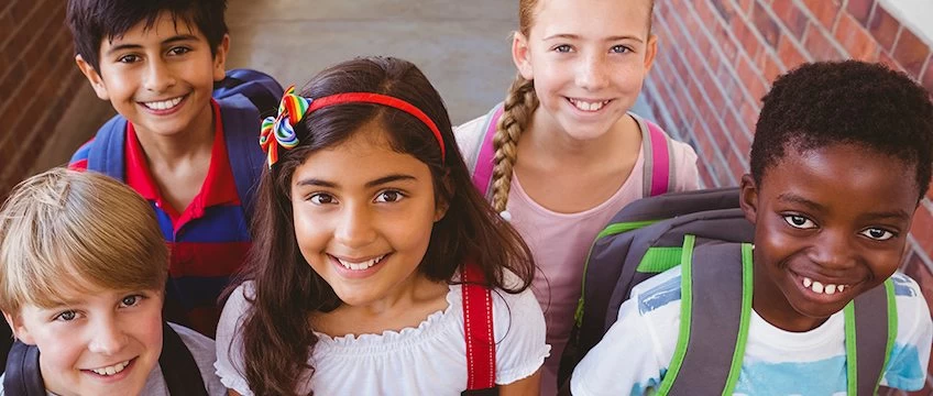 A group of children smiling with backpacks in front of a school building.
