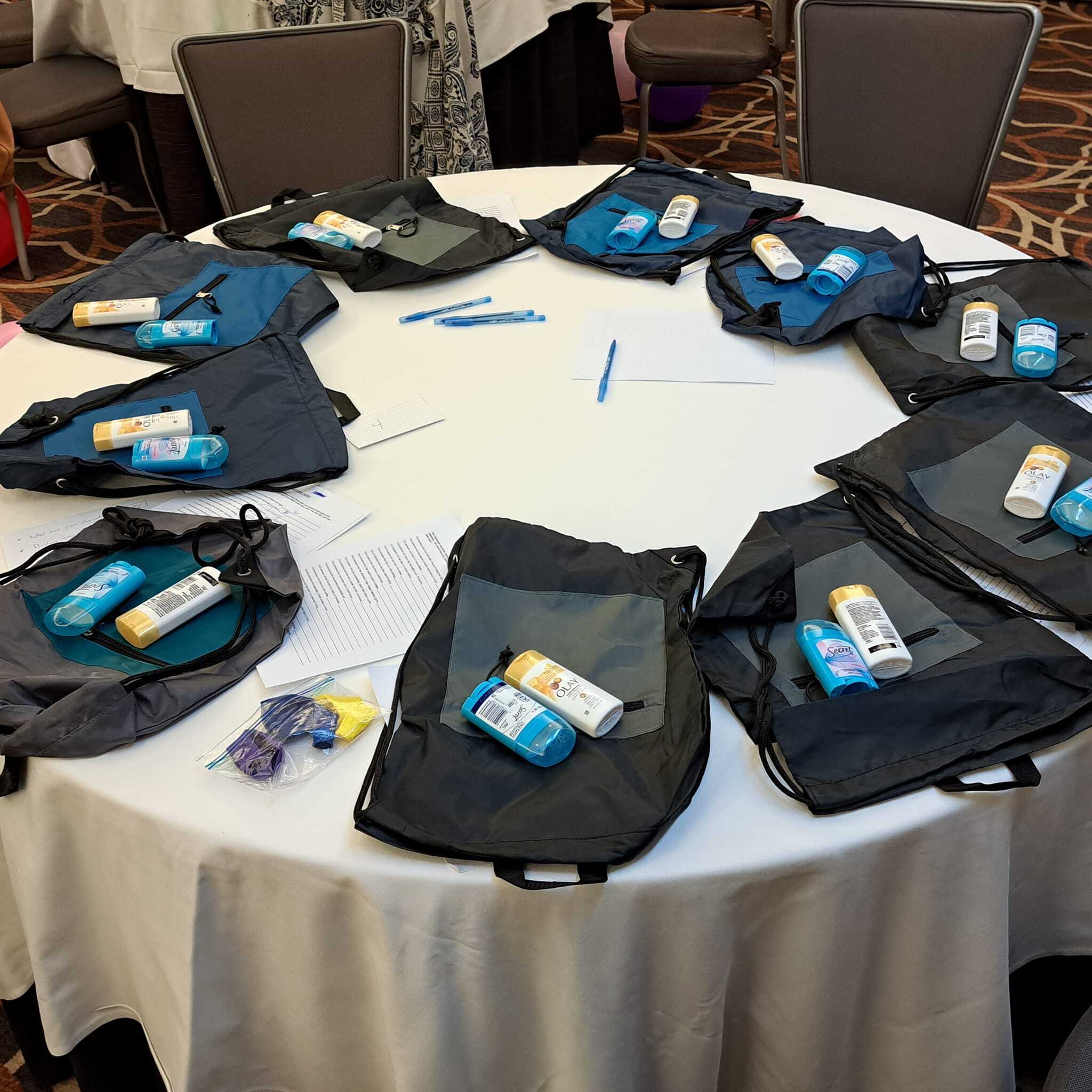 A round table with drawstring bags, sunscreen bottles, hand sanitizers, pens, and printed papers arranged at each seat.