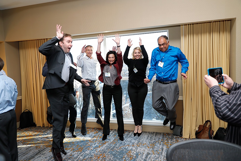 Five people in business attire are jumping in the air inside a conference room, while another person takes their photo with a smartphone.