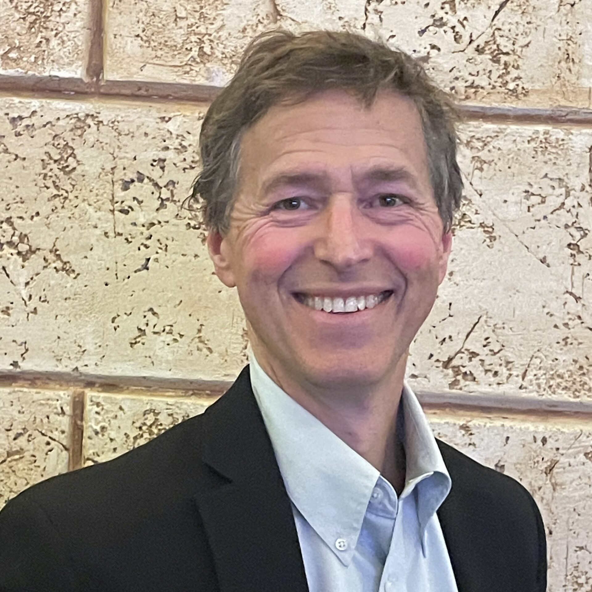 A man in a dark blazer and light blue shirt stands smiling in front of a textured beige wall.