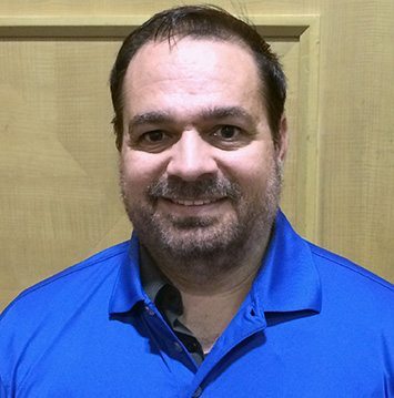 A man in a blue shirt smiling in front of a wooden wall.