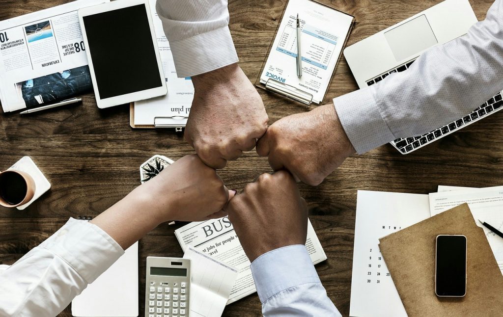 Four individuals fist-bumping over a business meeting table strewn with documents, electronic devices, and coffee cups.