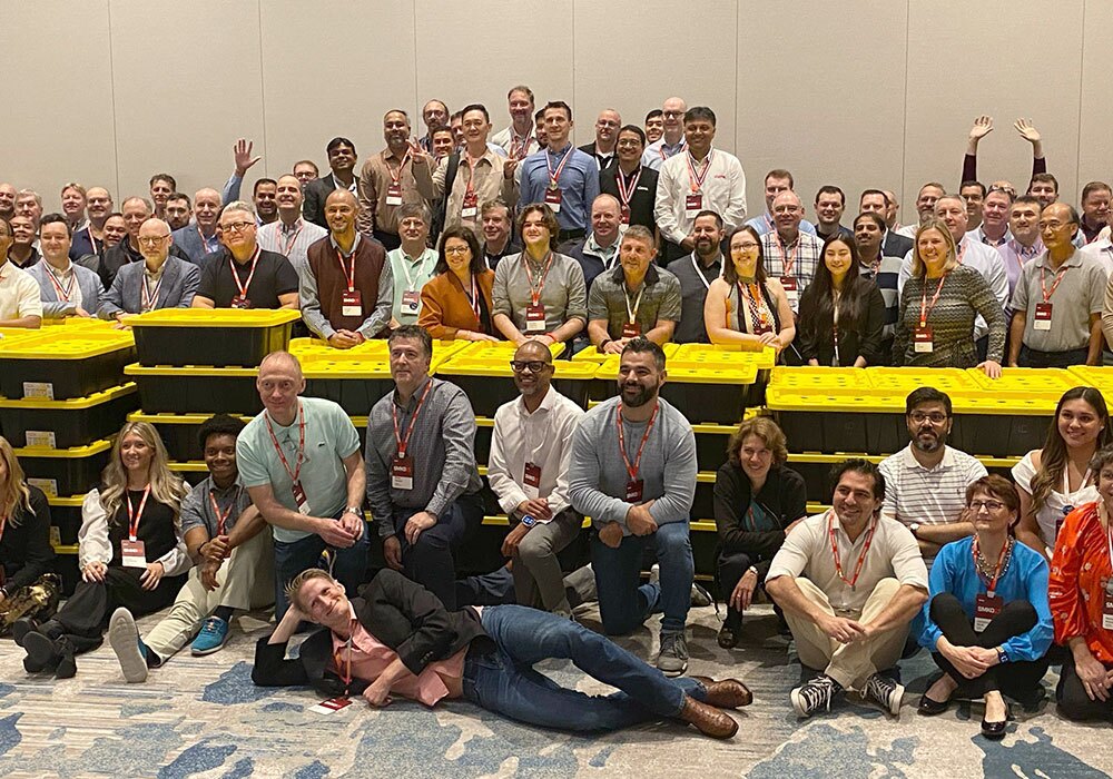 A large group of people poses for a group photo in front of yellow storage bins indoors, gathered after a successful hydroponics donation event. Some are standing, seated, or reclining in the front. Most wear name tags.