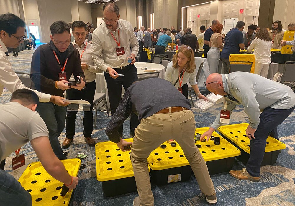 In a conference room, a group of people gathered around yellow containers with black lids, engaging in a lively discussion about hydroponics and exploring the potential impact of their upcoming donation.