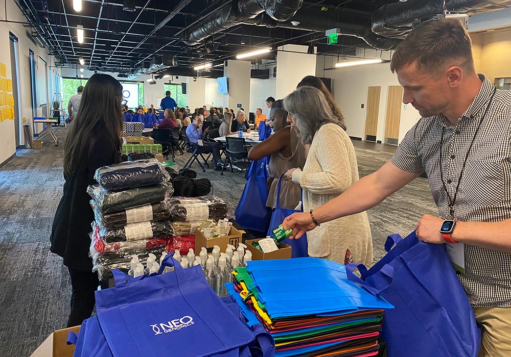 People are packing blue bags with various supplies, including blankets and hygiene products, in a large room set up for an event. More people are seated at tables in the background, preparing cancer care packs to provide comfort to those in need.
