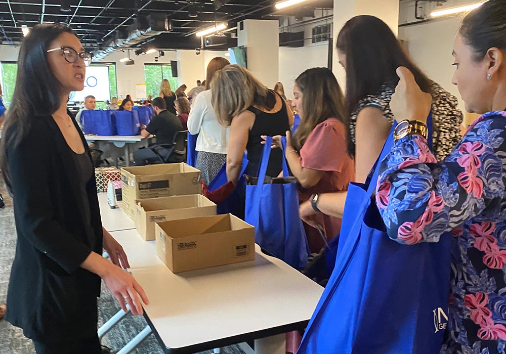 People stand in line to receive cancer care packs from cardboard boxes placed on tables in a large, well-lit room.