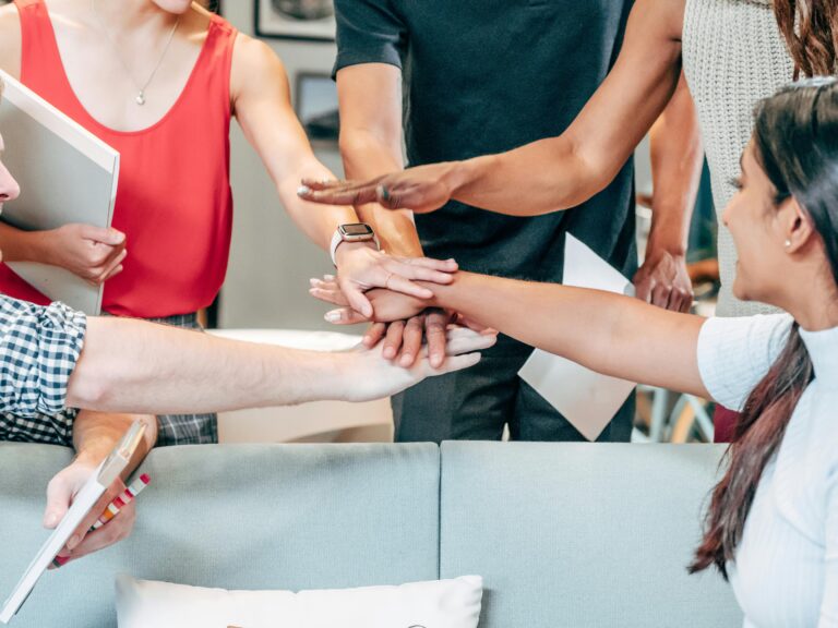 A group of people stack their hands together in a gesture of teamwork while seated around a sofa, with notebooks and papers visible as they discuss their professional development goals.