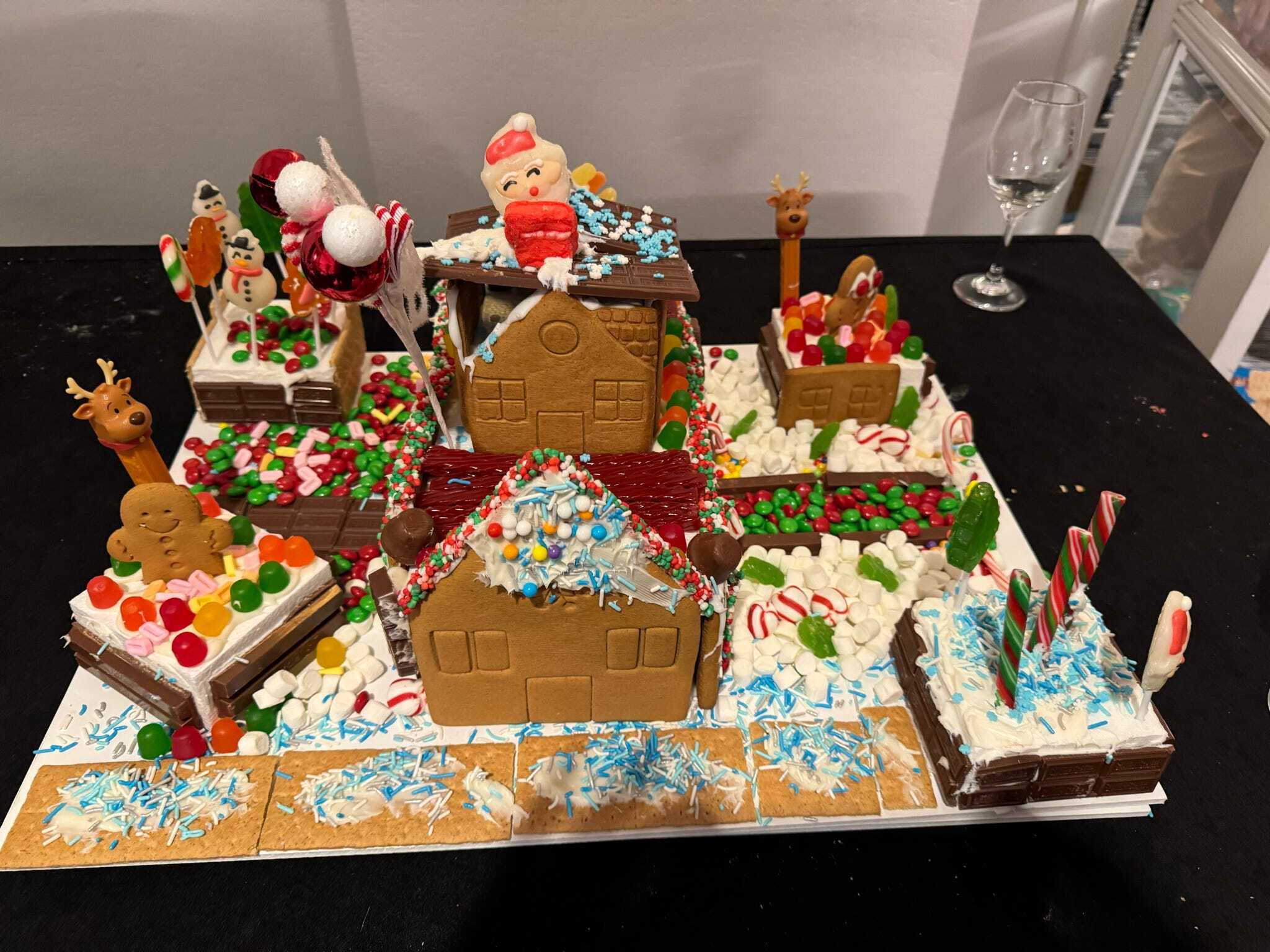 A festive gingerbread village scene with houses, candy decorations, Santa figure on the roof, reindeer, snow effects, and a wine glass in the background.