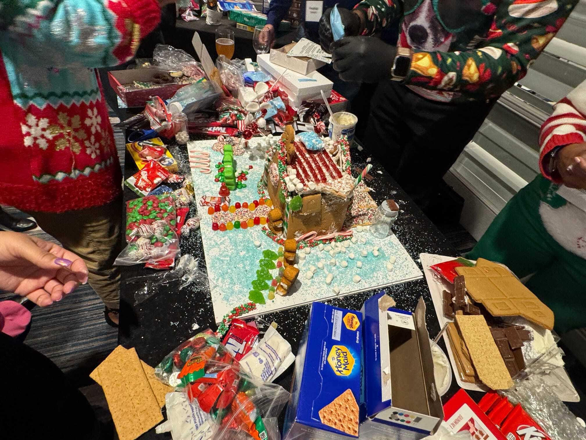 Several people are assembling and decorating a gingerbread house at a table covered with candy, icing, graham crackers, and holiday-themed supplies.