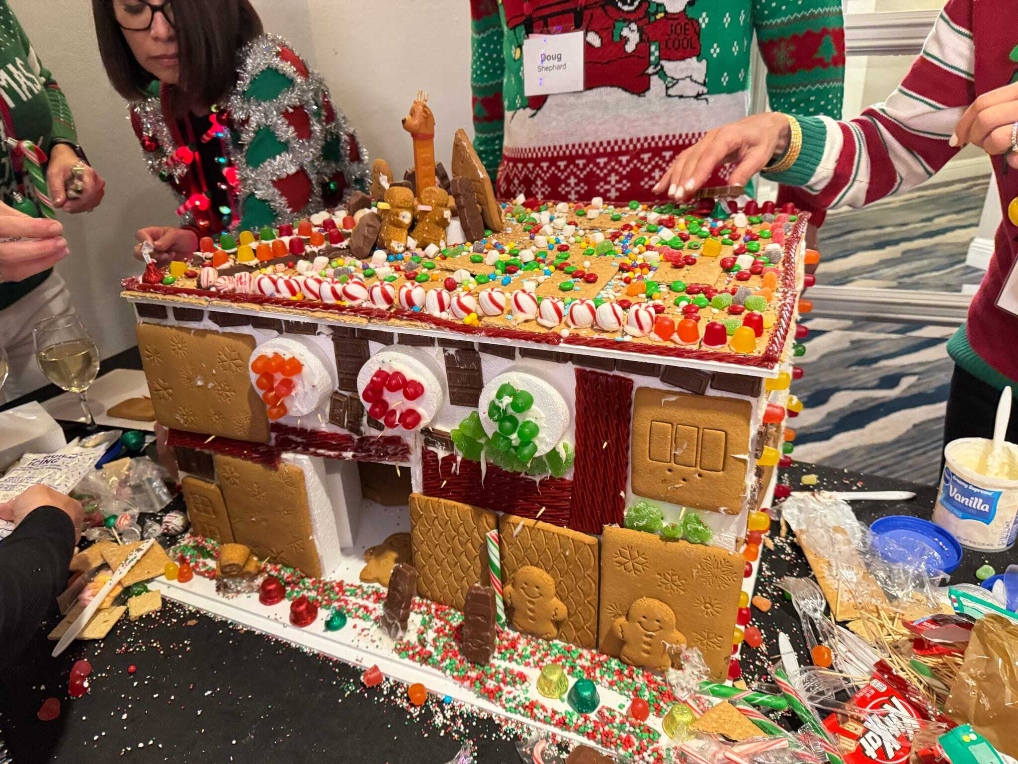 People decorate a large gingerbread house with candy, icing, and cookies. Two people wear festive holiday sweaters and there are decorating supplies on the table.