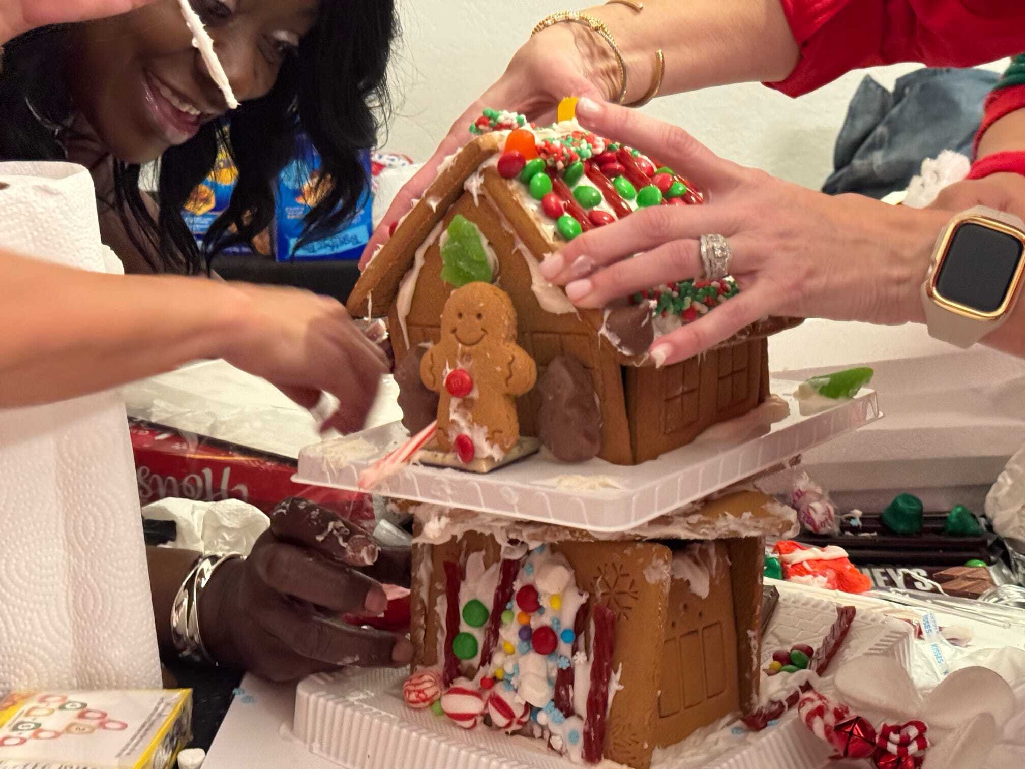 Three people are assembling and decorating a two-story gingerbread house with icing and colorful candies. One person holds a candy cane while another adjusts the roof.