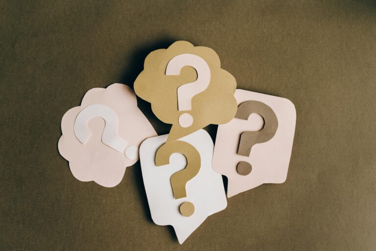 Four paper cutouts with question marks are arranged on a brown surface, each shaped like a speech or thought bubble in beige and light pink tones—perfect for sparking a question of the day for work.
