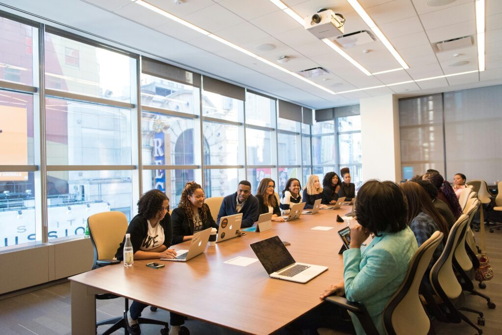 A group of people sit around a large conference table with laptops open, attending a meeting in a modern office with large windows.