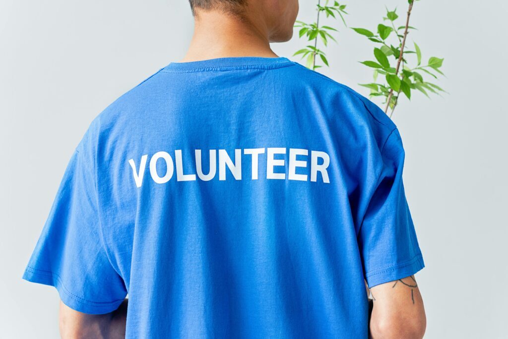 Person wearing a blue T-shirt with the word "VOLUNTEER" printed on the back, standing near a leafy green plant.