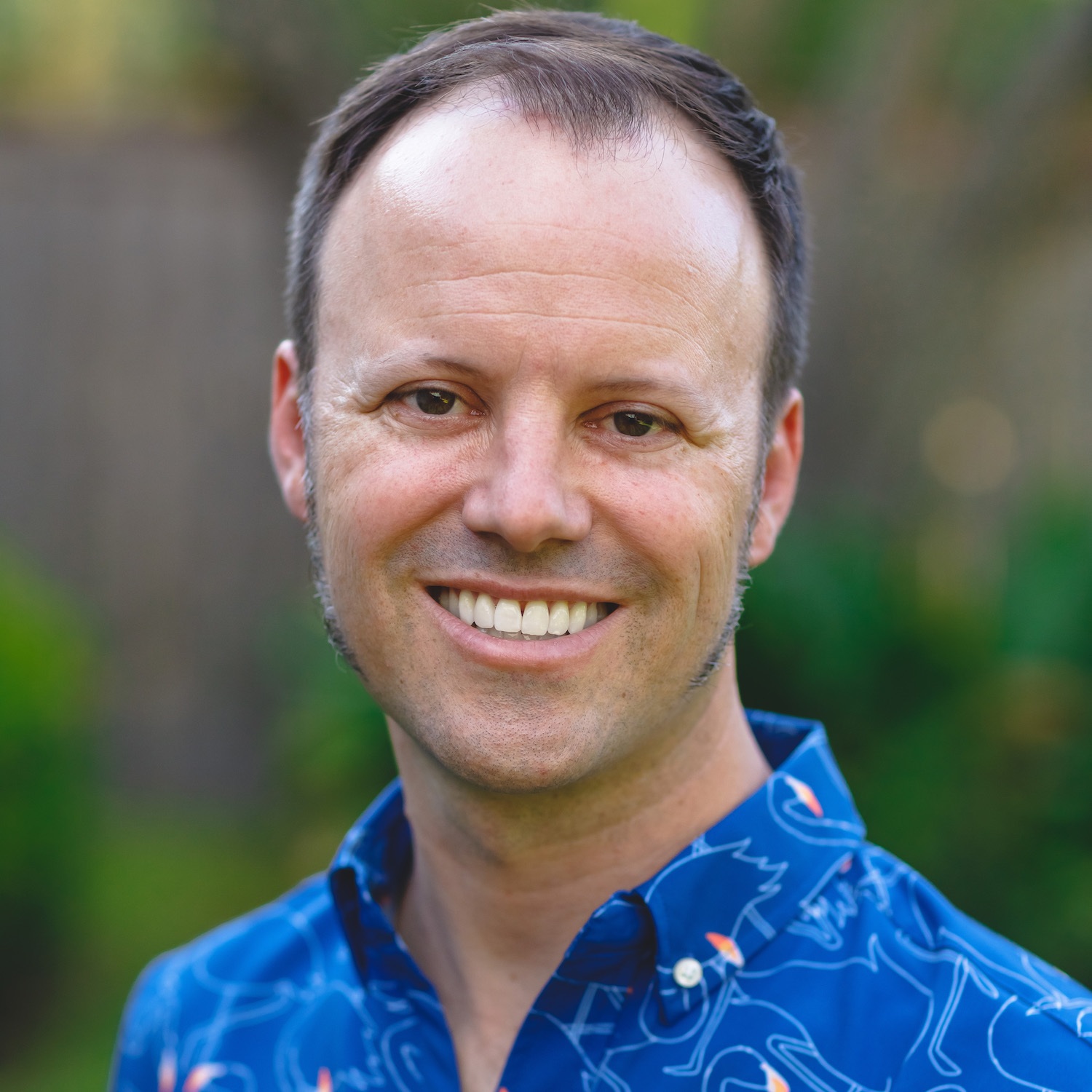 A man with short brown hair and sideburns smiles at the camera outdoors, wearing a blue button-up shirt with a colorful line pattern.