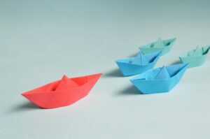 Five paper boats, one red and four blue, are arranged on a light blue surface. The red boat is positioned ahead of the blue boats.