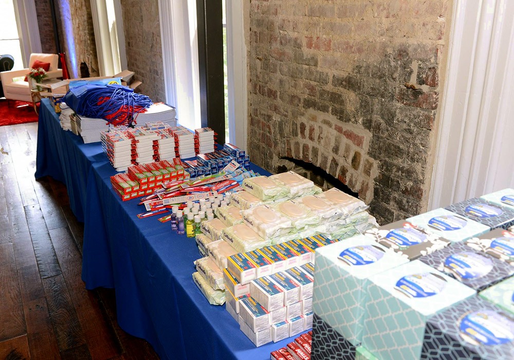 A table displays neatly arranged stacks of supplies, including towels, sanitary pads, toothpaste, soap, and tissue boxes, set against a brick wall backdrop.