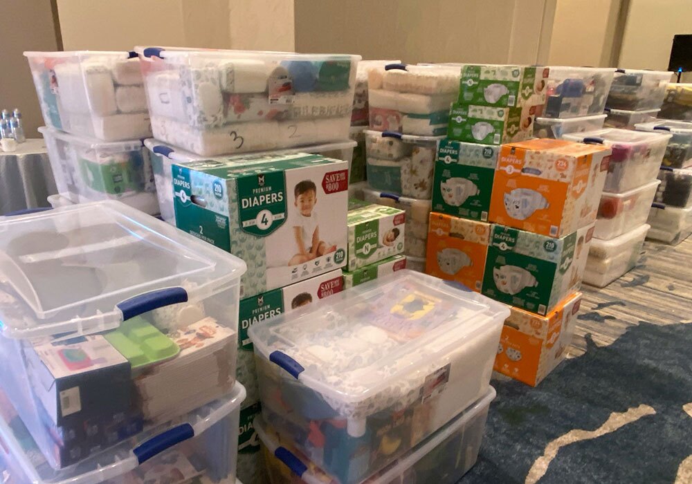 Stacks of diaper boxes and clear plastic containers filled with supplies are arranged in a room.