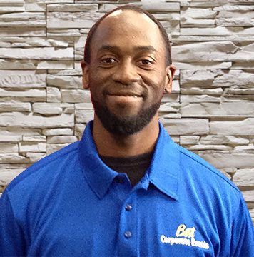 A black man in a blue shirt standing in front of a stone wall.