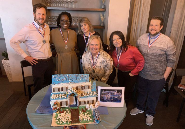 A group of people posing in front of a gingerbread house.
