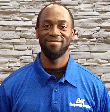 A black man in a blue shirt standing in front of a stone wall.