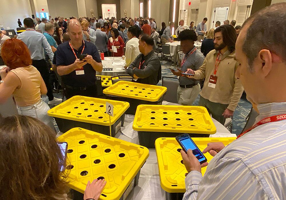 People gather around tables with yellow-lidded black containers, some looking at their phones in a busy conference room, as discussions about hydroponics donations fill the air.