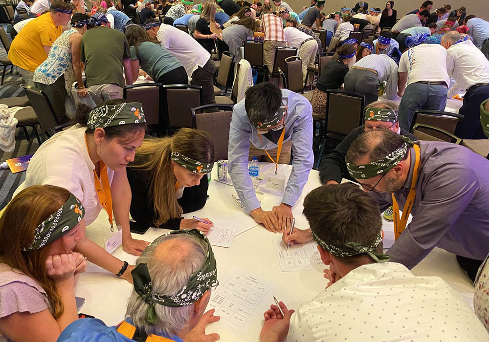 A group of people wearing patterned headbands gather around a table, engaged in a collaborative activity involving papers and pens. Other groups are visible in the background, participating similarly.