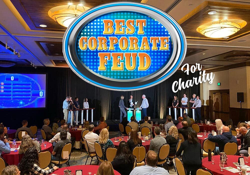 People competing in a corporate-themed game show event for charity, with an audience watching. A sign above reads "Best Corporate Feud.