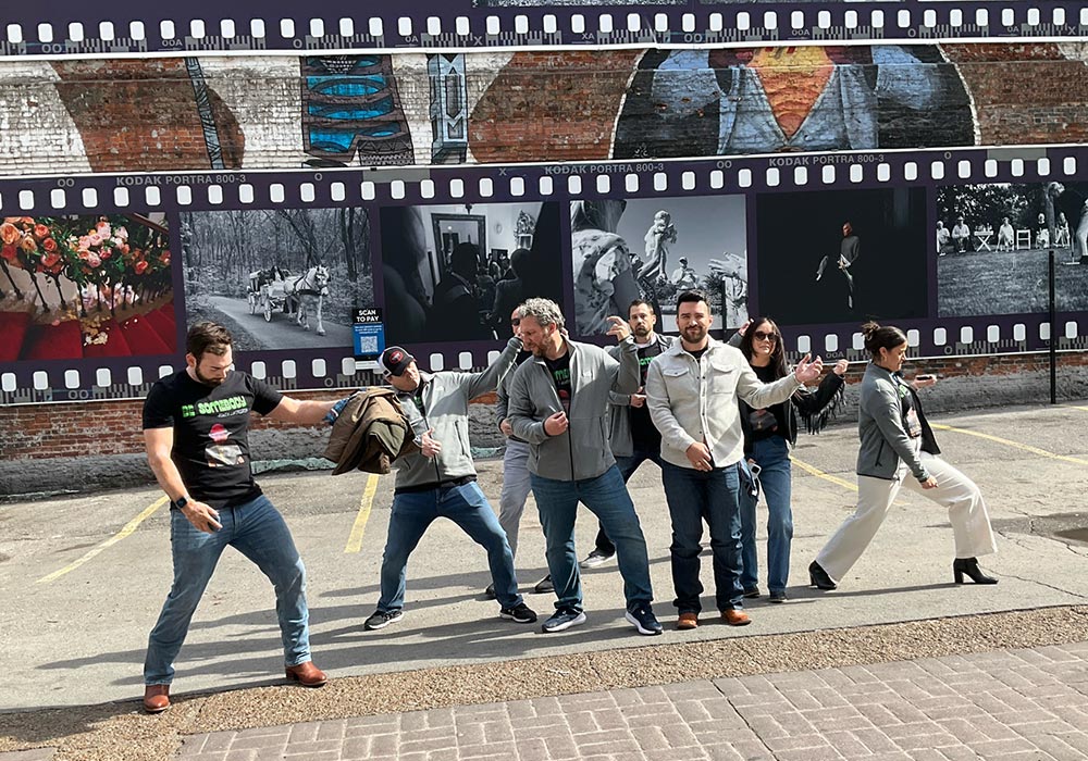 A group of people imitating the walking pose from the iconic beatles abbey road album cover in front of a large photo montage.