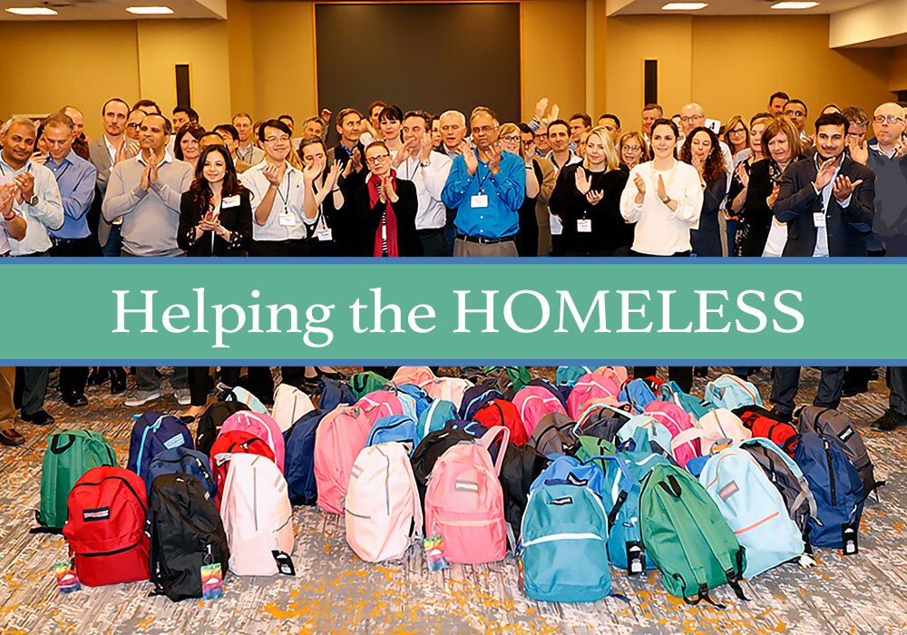 A group of people standing behind a collection of backpacks on the floor, with a banner reading "Helping the HOMELESS" in front.