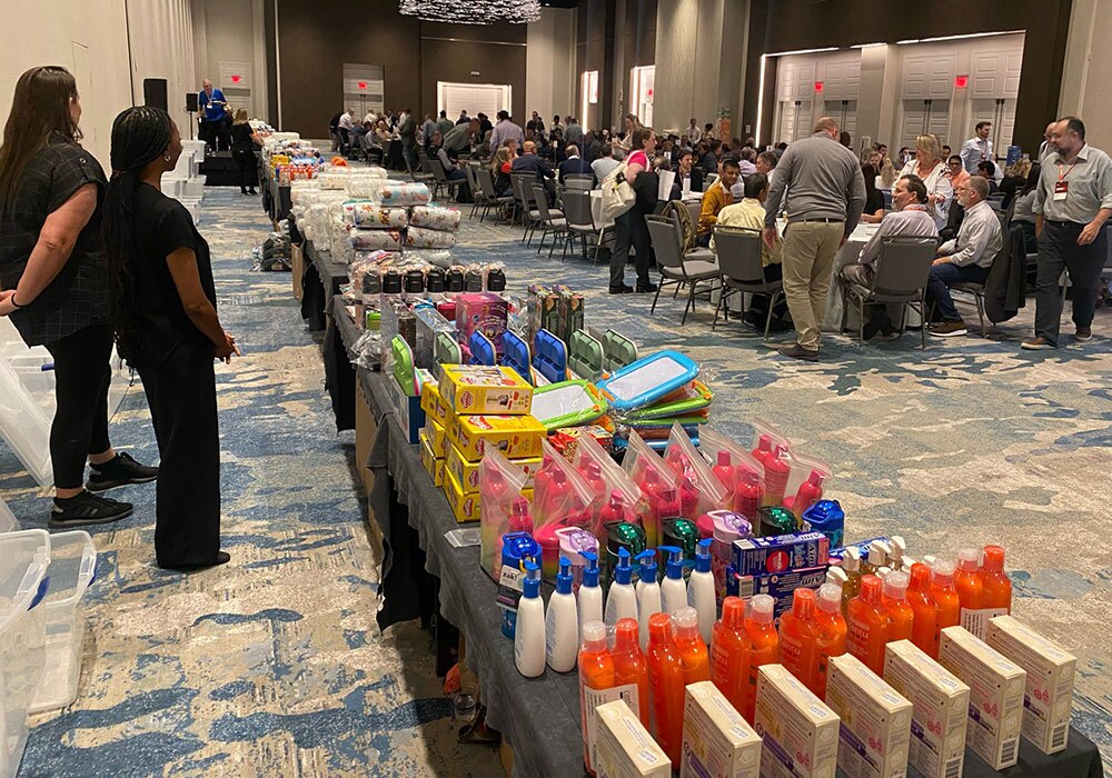 Room with tables filled with assorted toiletries and supplies, with people gathered at tables in the background.