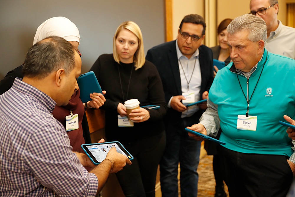 A group of people with name tags gather around a man demonstrating something on a tablet, while others hold tablets and observe attentively.