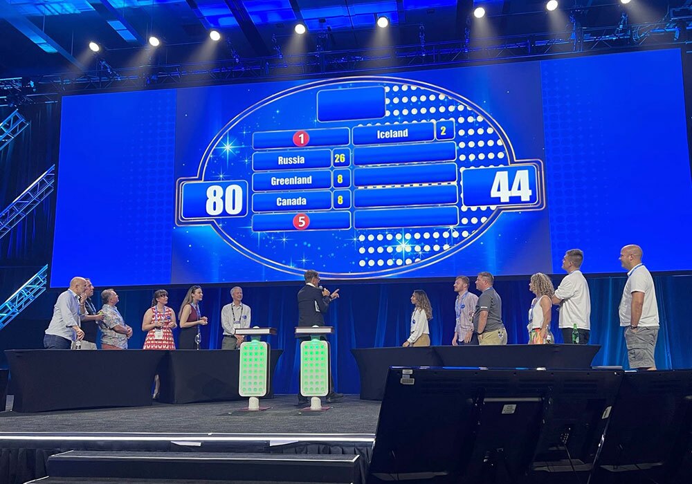 Two teams compete in a live Family Feud game on stage, with a host at the center and scores of 80 and 44 displayed on a large screen behind them.