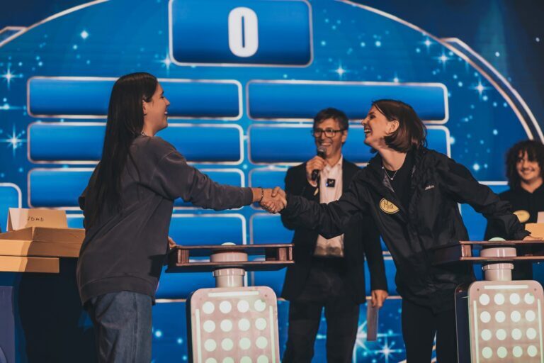 Two contestants shake hands at a game show podium while the host stands in the background, holding a microphone and smiling—perfect inspiration for Family Feud games for work.