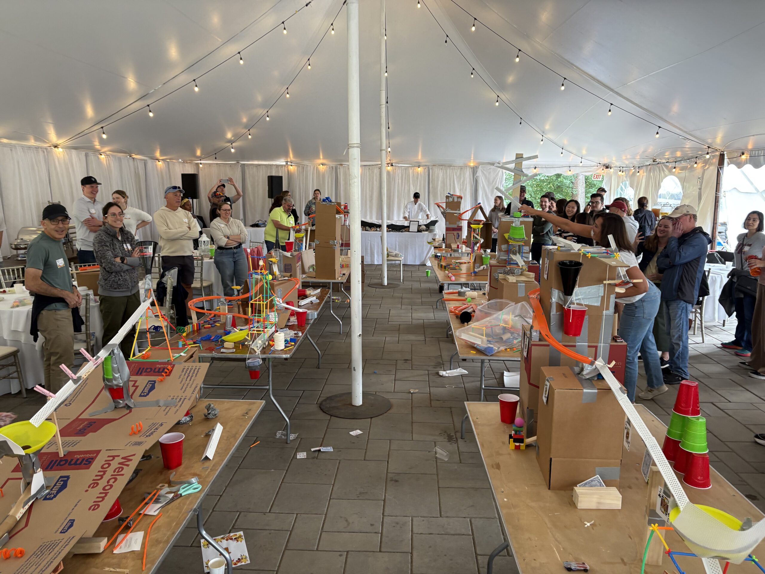 People stand under a large tent observing tables with various homemade Rube Goldberg machines made from cardboard, plastic cups, and other household materials.