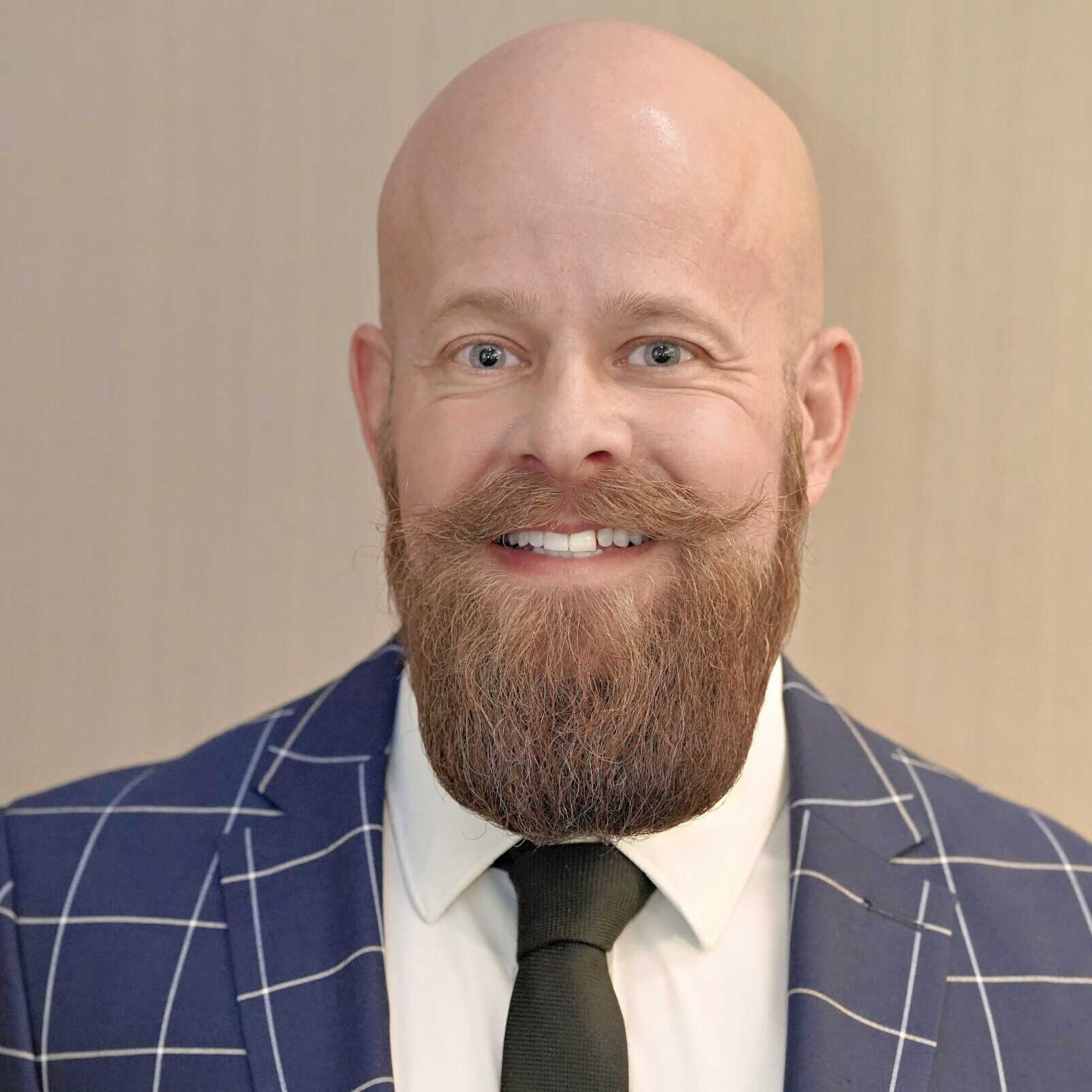 A bald man with a reddish-brown beard and mustache is wearing a blue checkered suit, white shirt, and black tie, standing against a beige background.