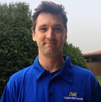 A man in a blue shirt standing in front of bushes.