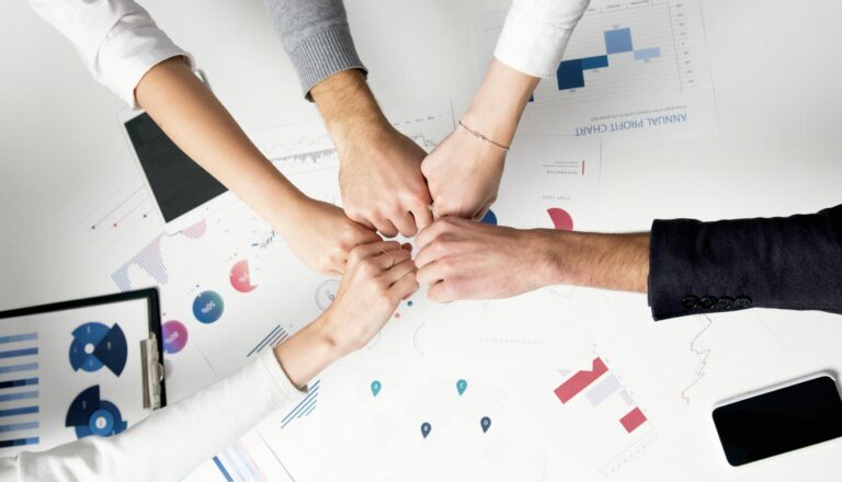 Five people do a group fist bump over a table with charts, graphs, and documents, symbolizing teamwork as they rebuild after employee layoffs in a business meeting.