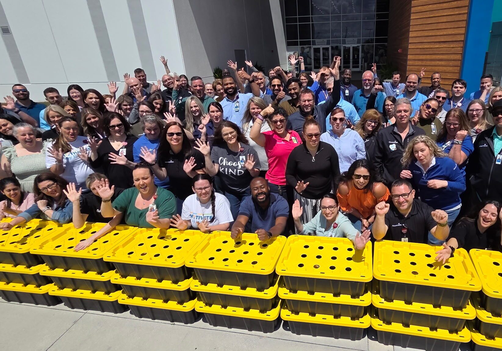 A large group of people standing and waving outside in front of several stacked yellow and black storage bins, near a building entrance during eco friendly team building activities.