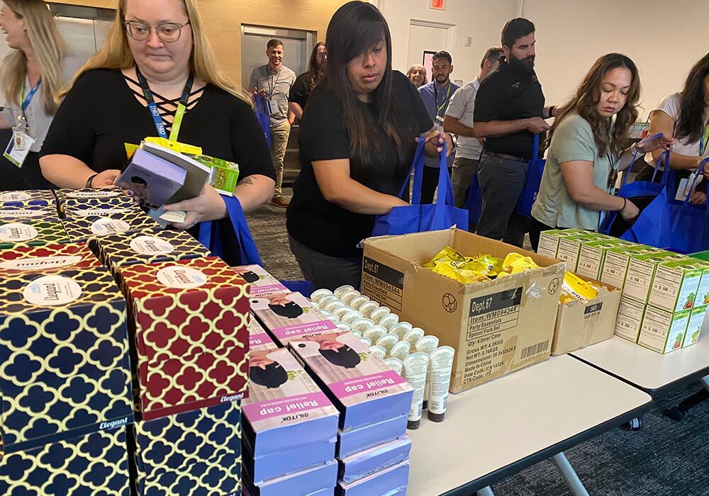 A group of people gathers supplies from a table loaded with various boxed items, placing them into blue cancer care packs.