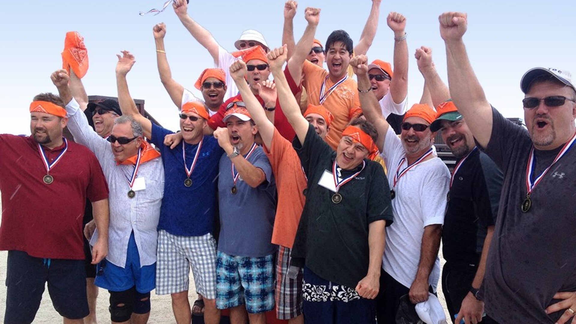 A group of men wearing orange bandanas and medals pose outdoors with raised arms, celebrating together.