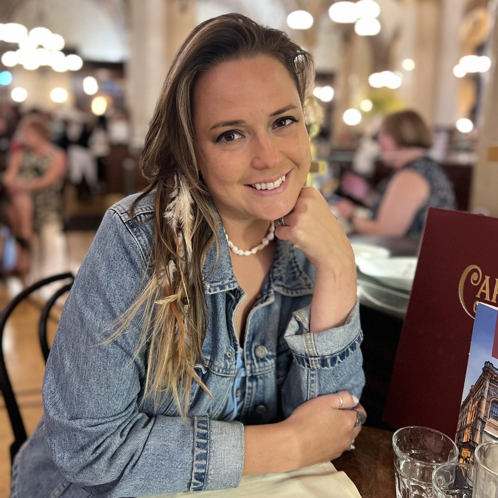 A woman in a denim jacket sits at a restaurant table, smiling at the camera. The background features arched ceilings and hanging lights, with other diners visible.