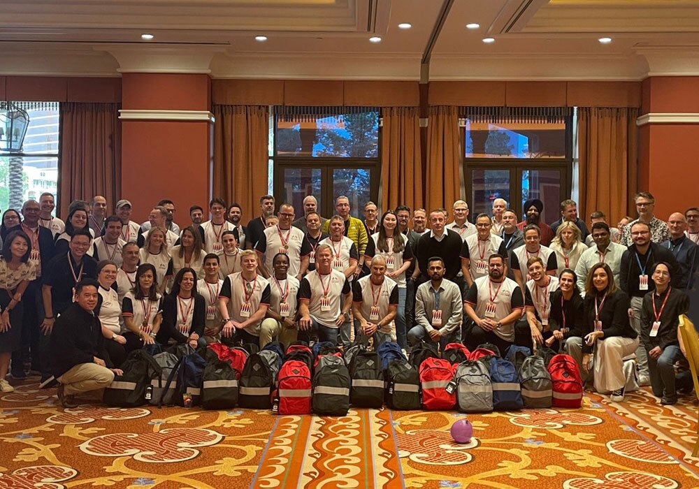A group of people posing for a photo in a large room with bags lined up on the floor in front of them. The room has patterned carpet and large windows in the background.