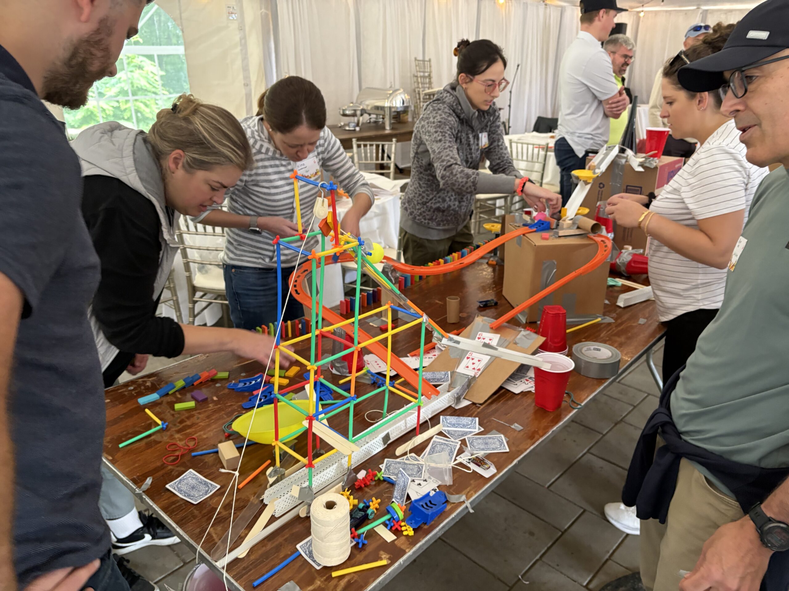 People gather around a table, collaborating to build a colorful, complex contraption using various materials like straws, cups, string, and playing cards.