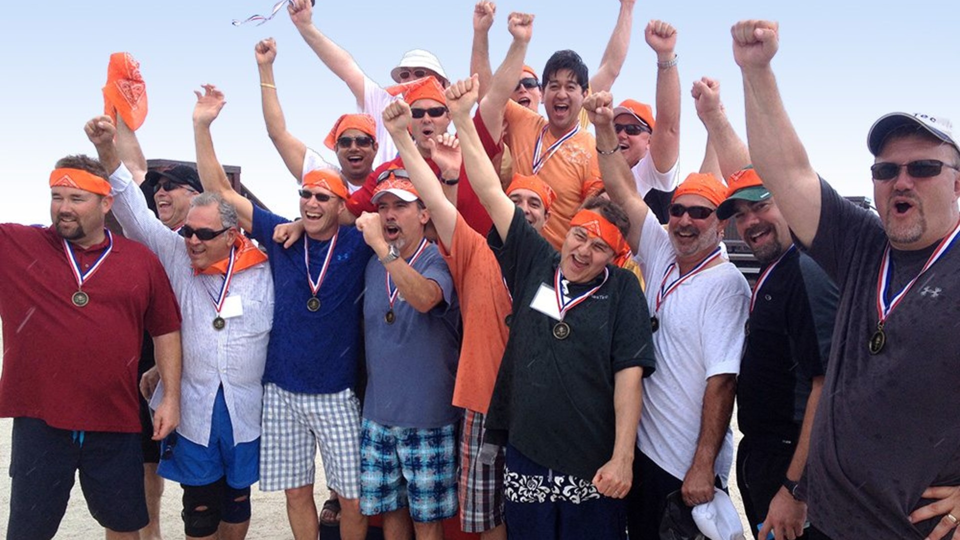 A group of men wearing orange bandanas and medals pose outdoors with raised arms, celebrating together.