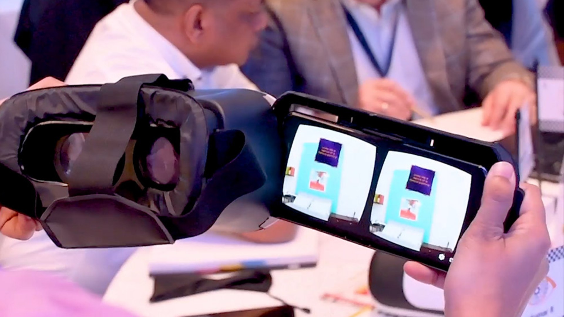 A person holds a VR headset with a smartphone displaying a virtual reality interface; three people are seated in the background.